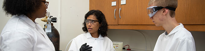 Three individuals in lab coats and goggles talking in a laboratory.