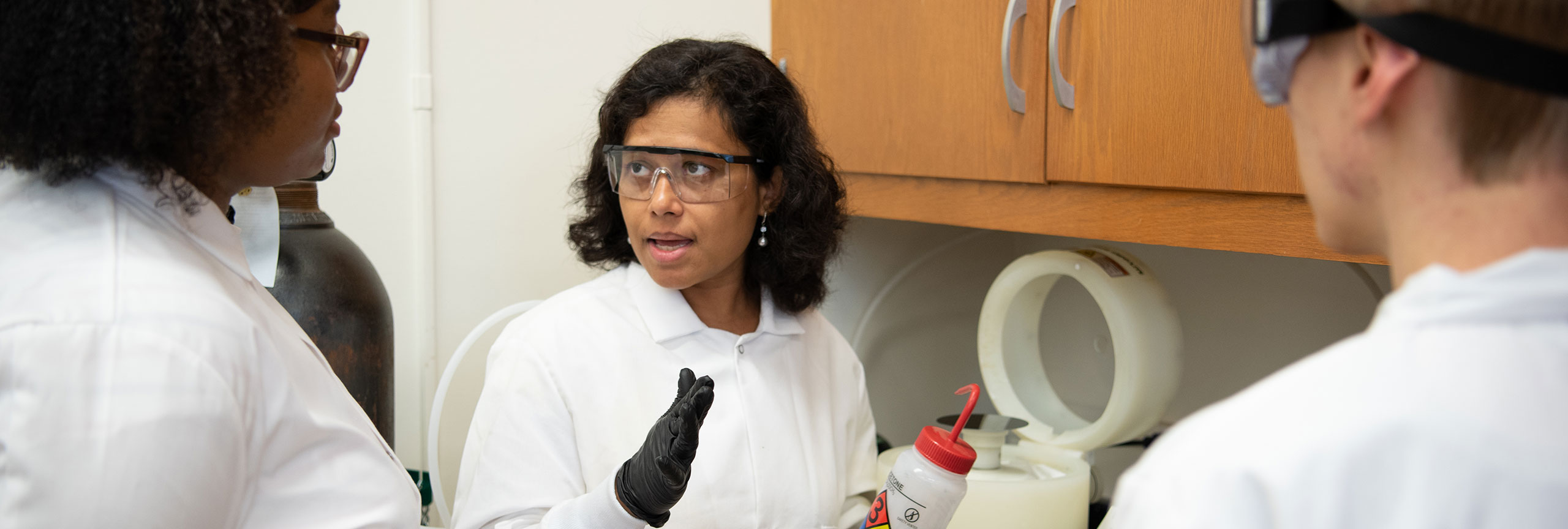 Three individuals in lab coats engage in conversation at a laboratory counter filled with equipment and supplies.