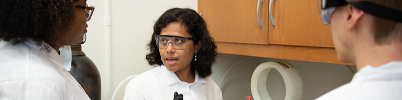 Three individuals in lab coats engage in conversation at a laboratory counter filled with equipment and supplies.